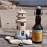 Bügelverschluss-Bierflasche neben Ton-Leuchtturm auf Holztisch, im Hintergrund Strand und Meer